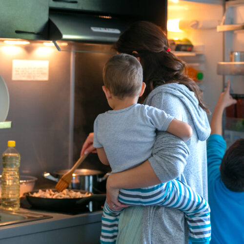 Mother carrying toddler while frying mushrooms for lunch, her older son pointing at stuff in the fridge